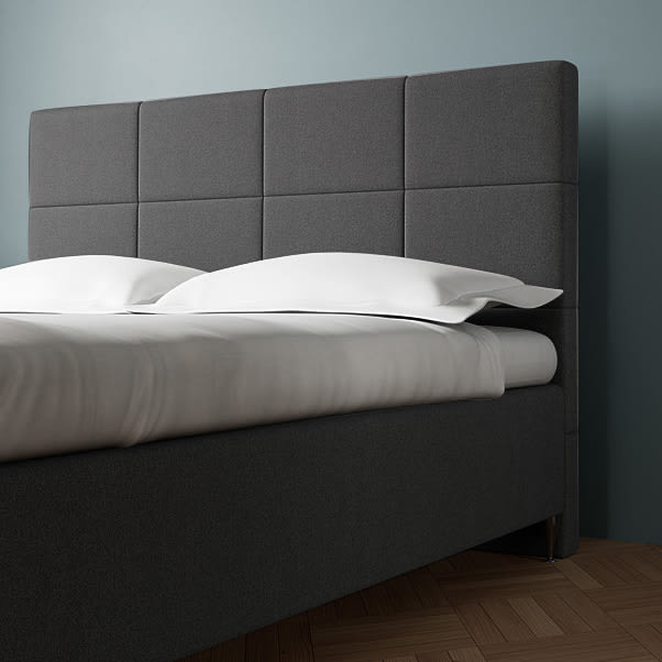 Mørkegrå stofbeklædt seng med et polstret hovedgærde med firkantede paneler og hvidt sengetøj.