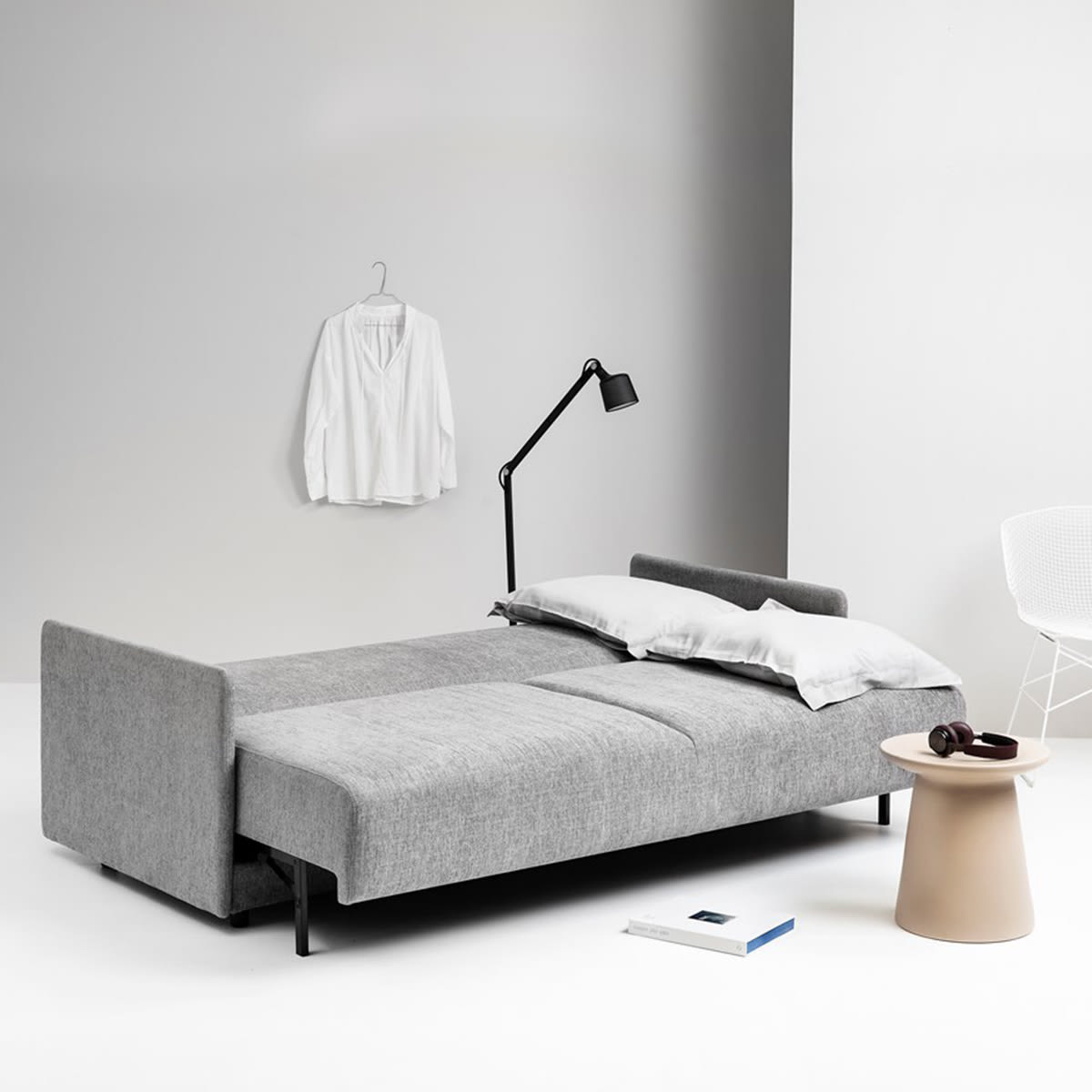 Grå sovesofa udslået til seng med hvide puder i et minimalistisk rum. En sort lampe og et lille bord står ved siden af.