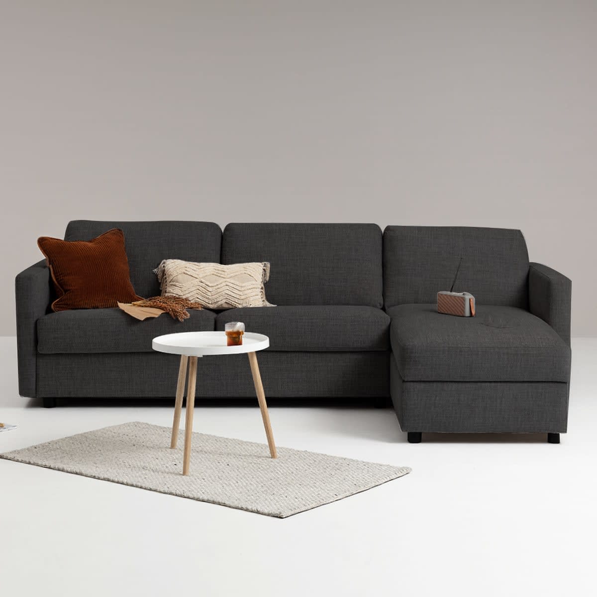 Mørkegrå L-formet sofa med pyntepuder, hvidt sidebord og tæppe. En radio ligger på chaiselongen.