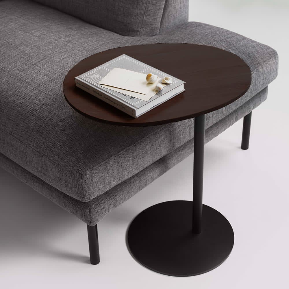 Grå stofsofa med et rundt mørkt træ sidebord. En bog og små dekorationer ligger på bordpladen.
