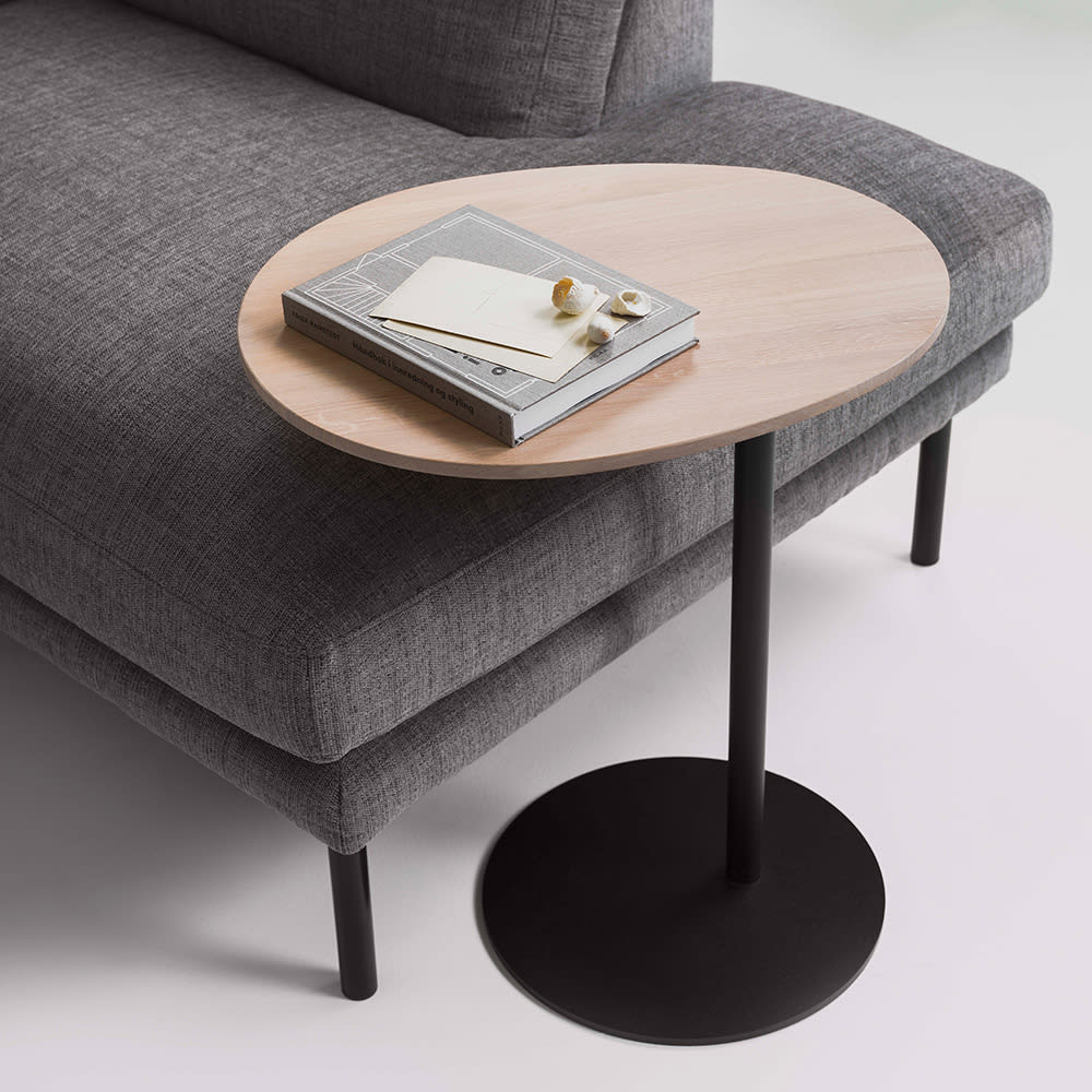 Moderne sidebord med oval bordplade i lyst træ og sort stel, placeret ved en grå sofa.