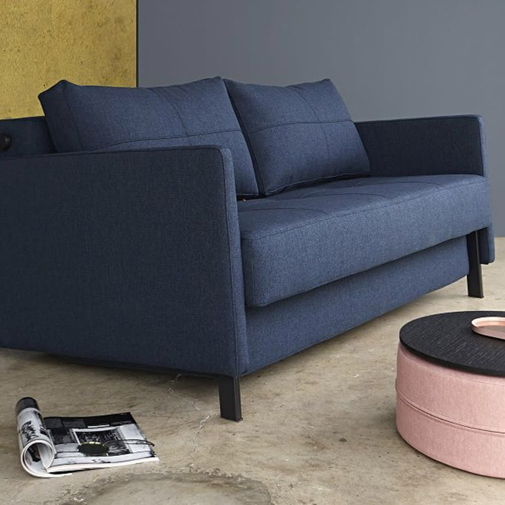 En mørkeblå stofsofa med sorte ben og firkantede syninger. En rosa puf står ved siden af.