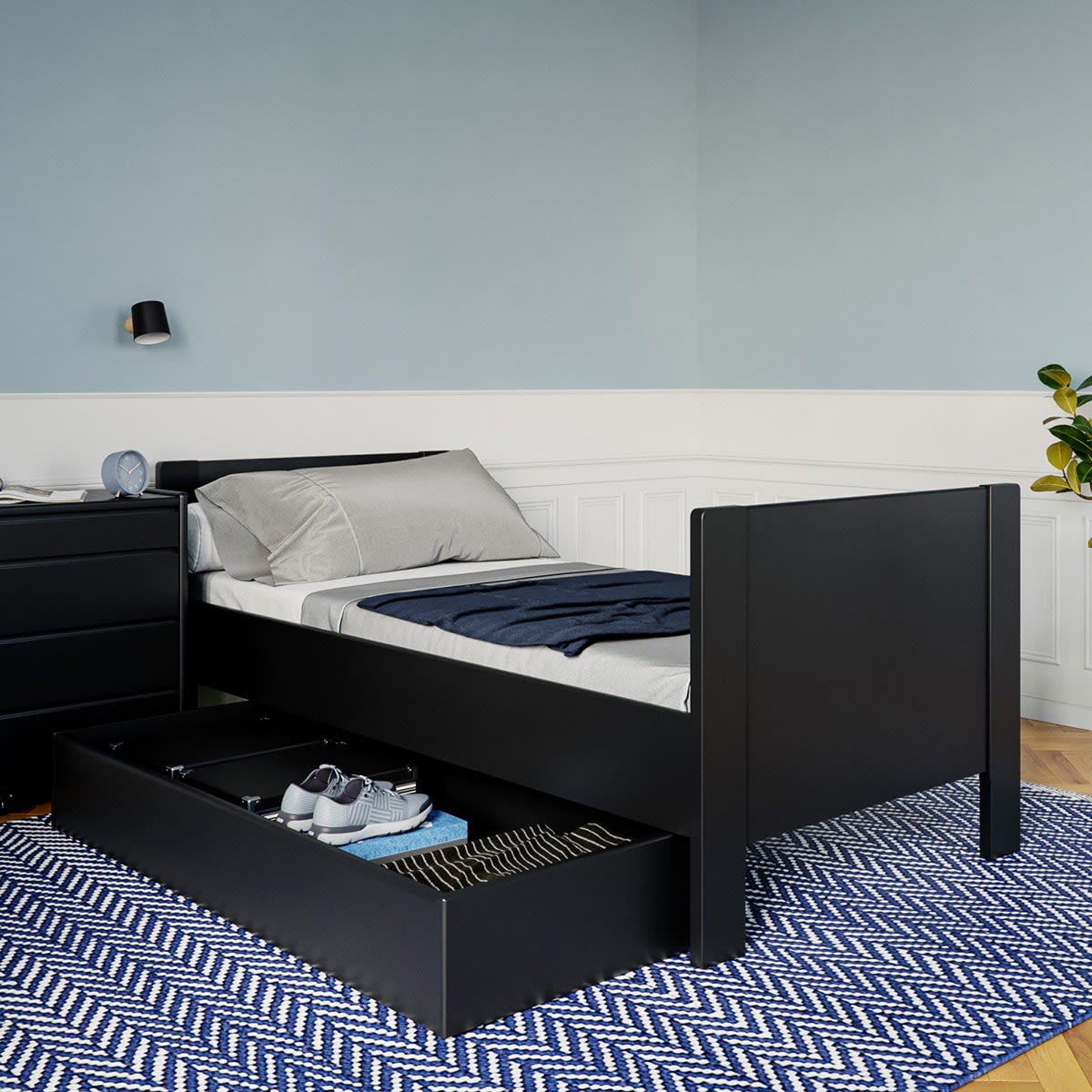 Sort enkeltseng med åben opbevaringsskuffe under, fyldt med sko. Sengen har gråt sengetøj og et blåt tæppe på et blåt-hvidt tæppe.