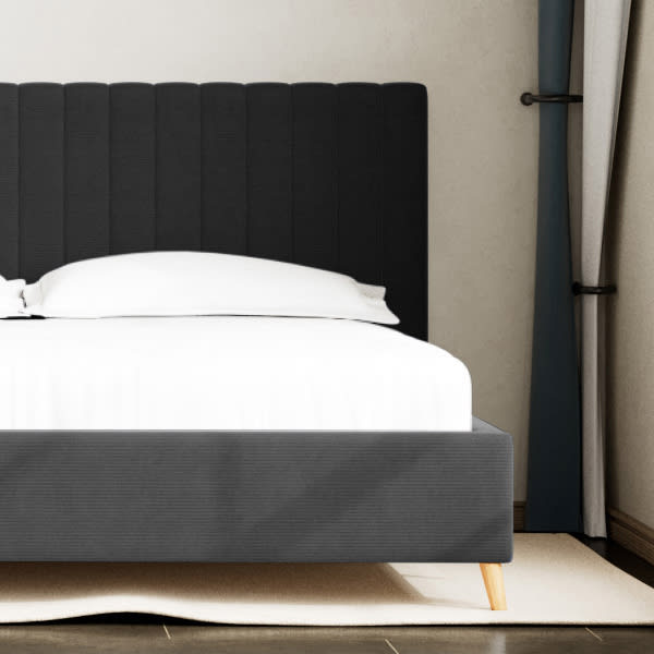 Et mørkegråt polstret sengestel med paneleret sengegavl og hvidt sengetøj. Sengen har lyse træben og står på et lyst tæppe.