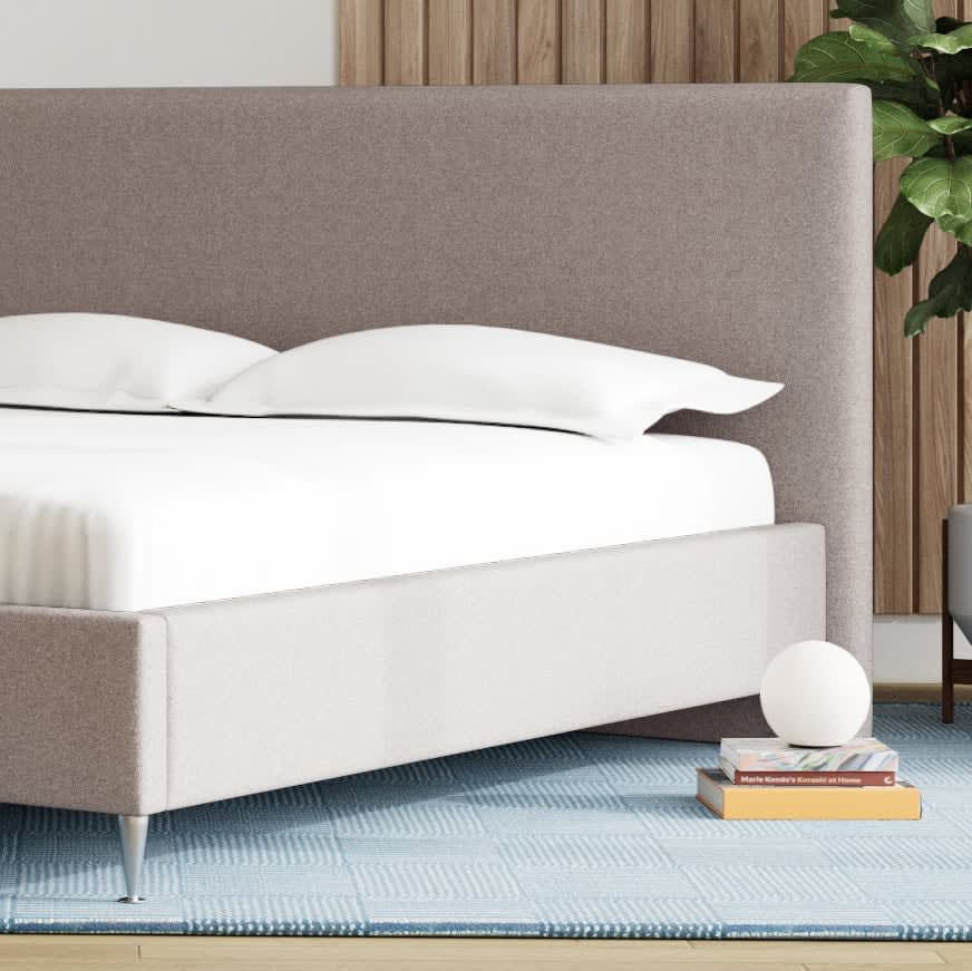 En gråbrun, polstret seng med hvidt sengetøj og et blåt mønstret tæppe.