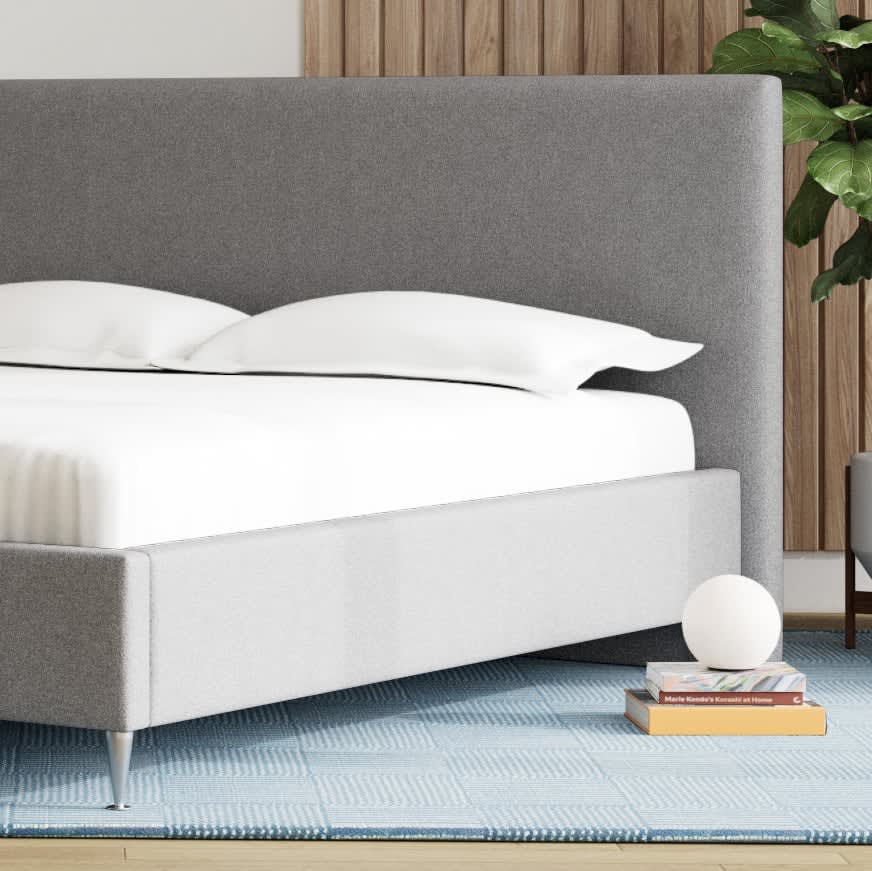 Moderne seng med gråt polstret sengegavl og lysegrå ramme, på et blåt mønstret tæppe.