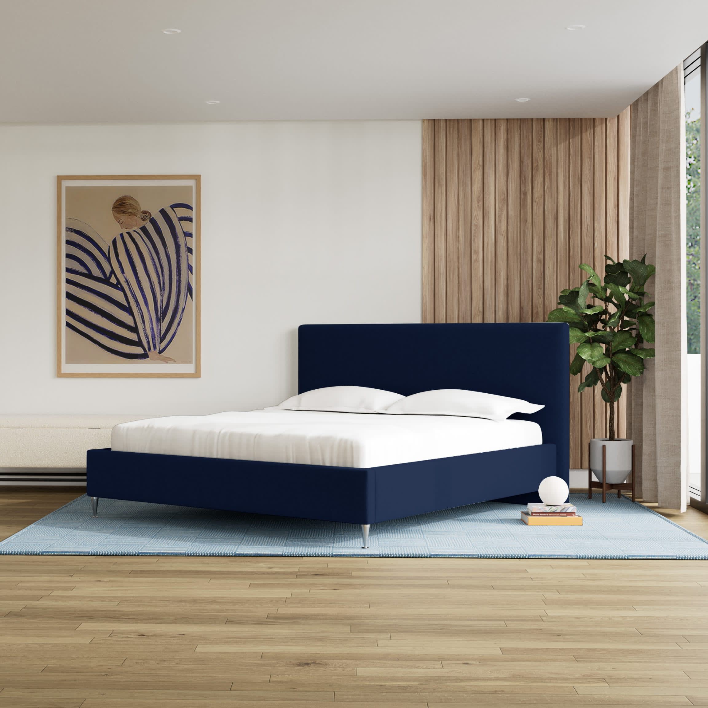 Et moderne soveværelse med en navyblå seng, hvidt sengetøj og et blåt tæppe på trægulvet. En illustration hænger på væggen.