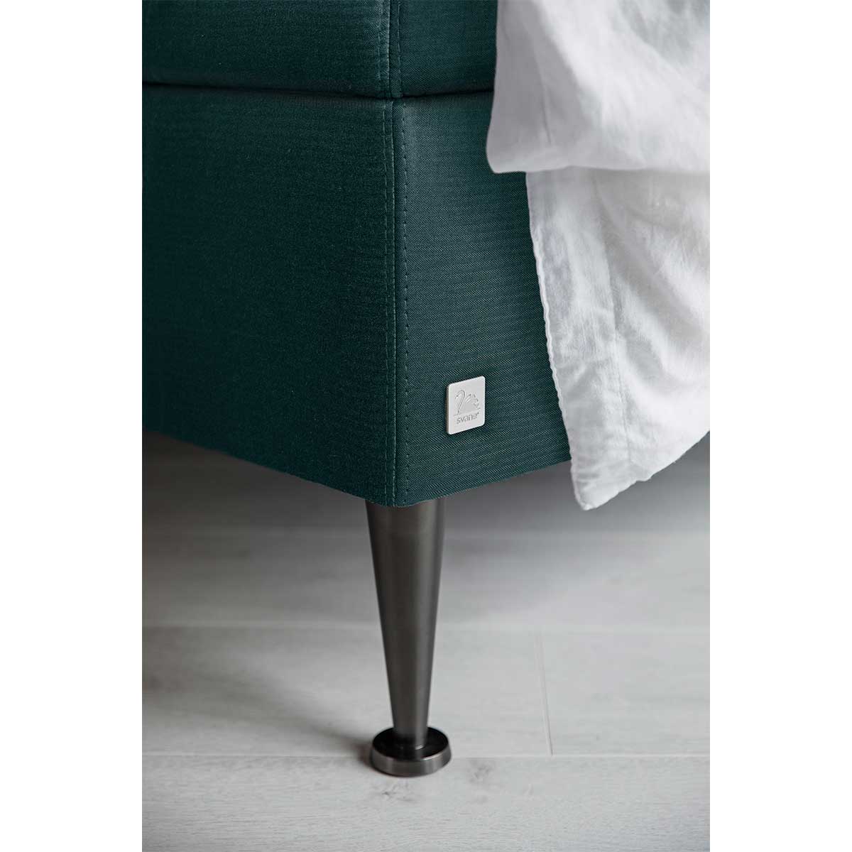 Nærbillede af mørkegrøn sengebund med en metalben og hvidt sengetøj. Svane-logoet ses på siden.