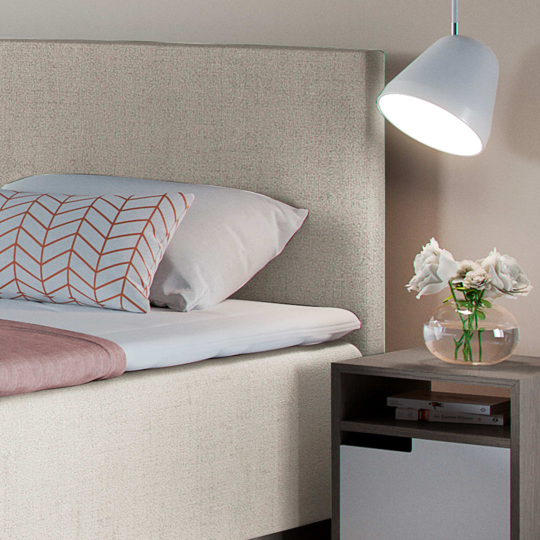 En seng med lysgrå sengegavl, hvidt sengetøj og en pude. En hvid pendellampe lyser op, og et sengebord har blomster.