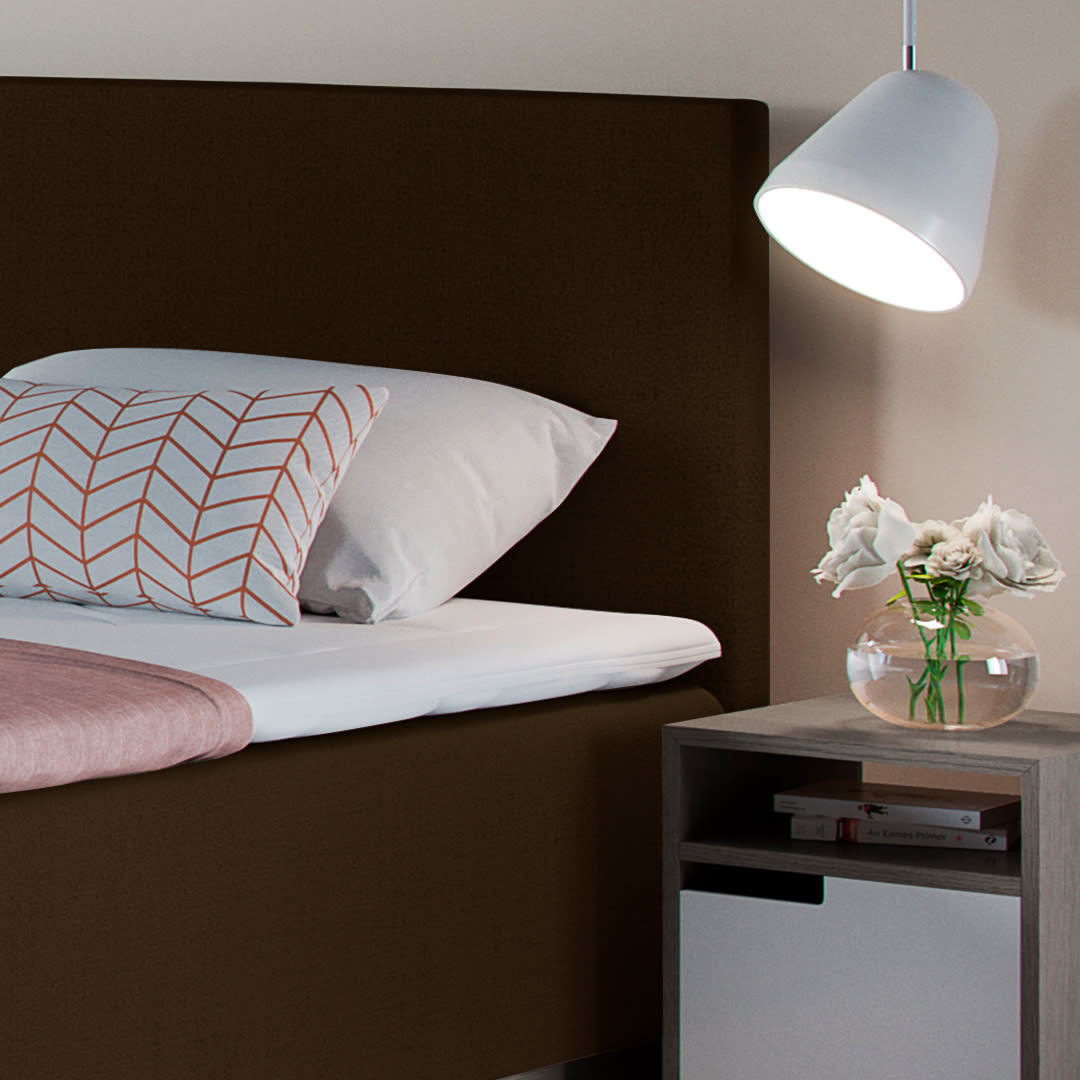 Et soveværelse med en brun seng, hvide puder og et lyserødt tæppe. Et sengebord med blomster og en hvid lampe.