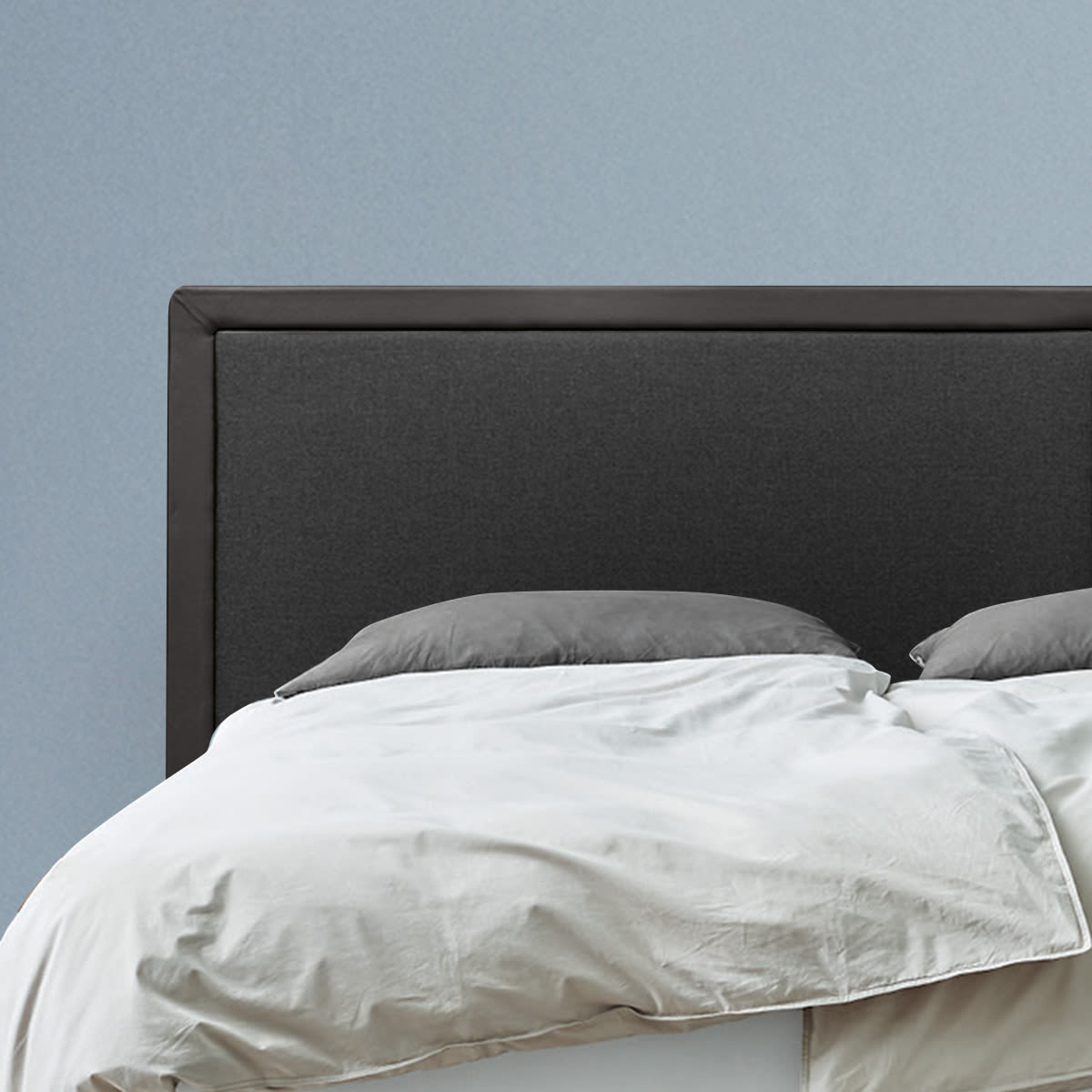 Seng med mørkegrå polstret sengegavl, lysegrå dyne og puder foran en lyseblå væg.