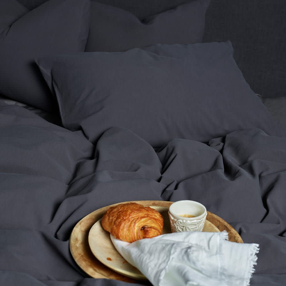 Mørkegråt sengesæt med en bakke på sengen. På bakken ligger en croissant, en lille kop kaffe og en hvid serviet.