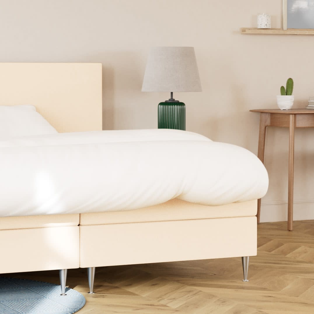 Lysbeige seng med hvidt sengetøj i et soveværelse. En mørkegrøn lampe og kaktus på natbordet.