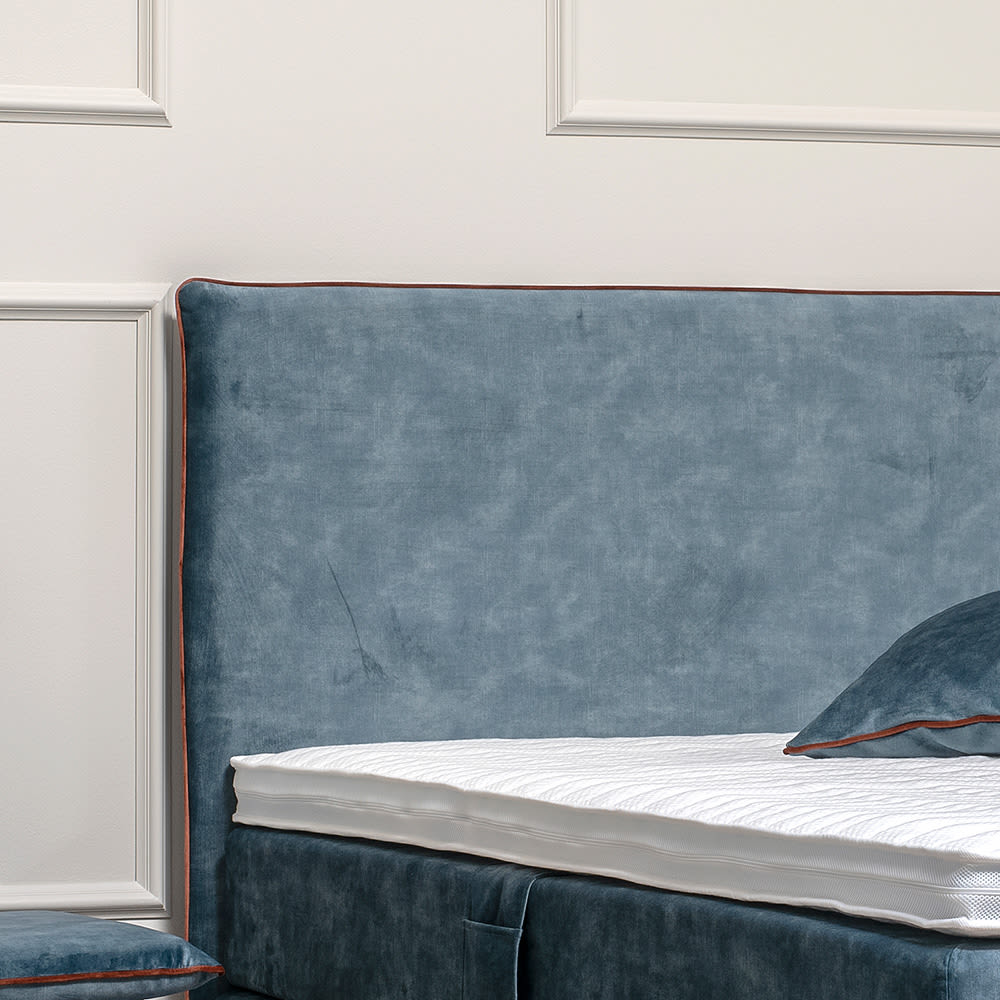 Blå velour hovedgavl med orange kant og matchende seng med hvid topmadras. Placeret foran en hvid væg med stuk.