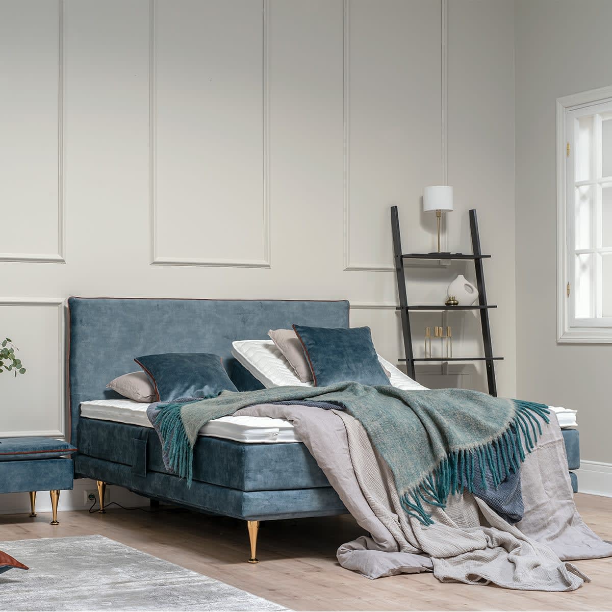 Elegant blå velour kontinentalseng med guld ben og matchende puf i et soveværelse med en stigehylde.
