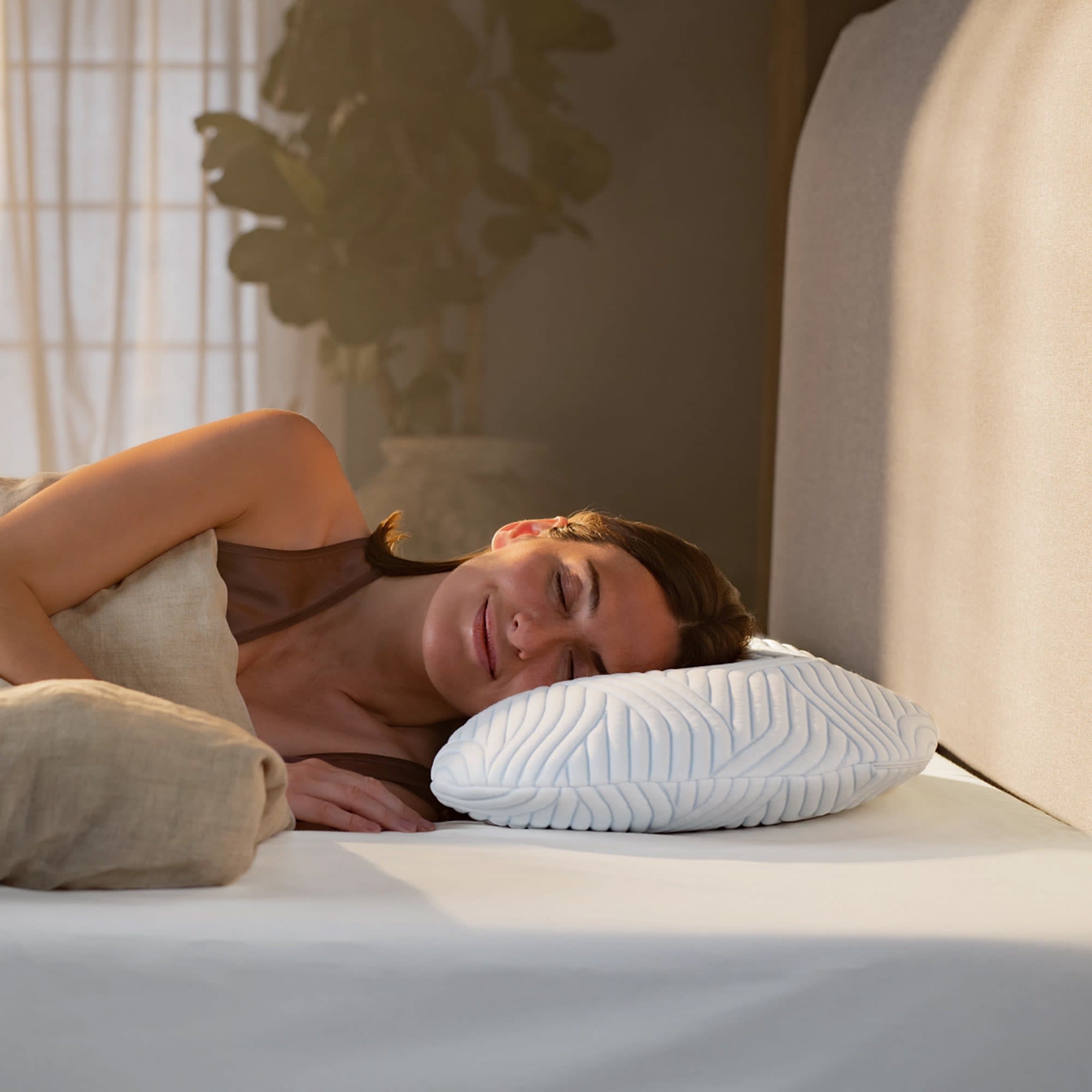 En kvinde sover fredeligt på siden i en seng med en hvid, tekstureret pude. Hun smiler let med lukkede øjne.