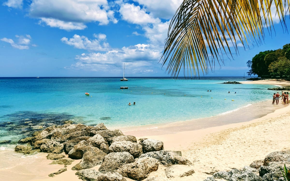 Turquoise water, a sandy shoreline and rocky foreground at Sandpiper Beach, Barbados, with swimmers and a sailboat offshore.