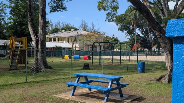 Fenced children’s playground with swings, climbing equipment and a blue picnic table at Folkestone Beach in St. James, Barbados.