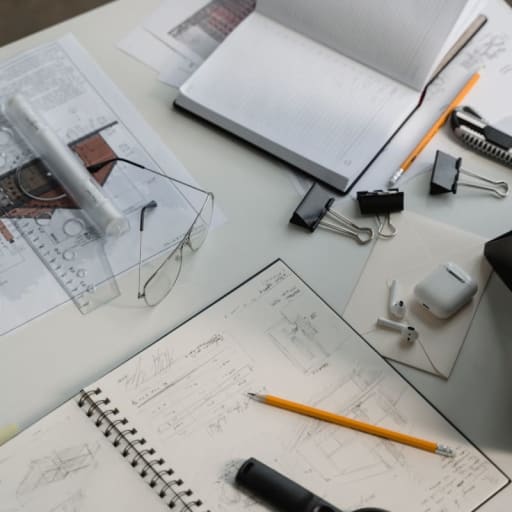 A desk strewn with paper notebooks, pencils, glasses, rulers and other architectural design tools
