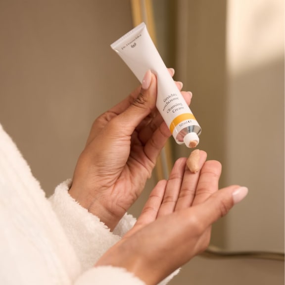 Hands squeezing cleansing cream from a tube onto a fingertip, with a soft beige bathroom background.