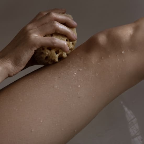 Close-up of a person scrubbing a wet arm with a natural sponge in the shower.