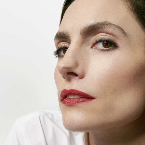 Close-up of a woman with smoky eye makeup, red lipstick, and a neutral expression against a white background.