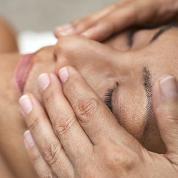 Close-up of a facial massage with hands gently pressing a person's closed eyes and cheeks.