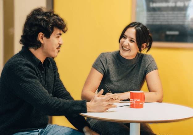Deux personnes discutant et souriant autour d'une petite table blanche. L'homme à gauche porte un pull noir, et la femme à droite porte une t-shirt gris et écris sur un carnet. Une tasse orange est posée sur la table, et le mur en arrière-plan est jaune