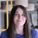 Portrait de Marie-Hélène, souriante, aux cheveux longs bruns, portant un haut violet. L'arrière-plan montre un environnement de bureau avec des éléments de décoration modernes