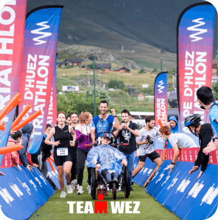 Photo d'une équipe en pleine course lors du Triathlon de l'Alpe d'Huez. Au centre, Théophile en fauteuil roulant est poussé par son coéquipier Valentin, souriants, entourés de coureurs. Bannières colorées et spectateurs encouragent la TEAM WEZ.