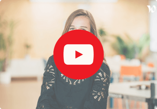 Lauriane portant une robe noire avec des motifs ajourés, assise dans un espace lumineux avec des plantes et des meubles colorés en arrière-plan. Un grand logo YouTube rouge est superposé au centre de l'image, suggérant un lien vers une vidéo.