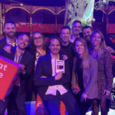 Photo de groupe lors de la cérémonie "Great Place to Work" : ils posent autour de cubes rouges portant le logo et un trophée. Décor de cirque avec éclairage coloré et ambiance joyeuse et conviviale.