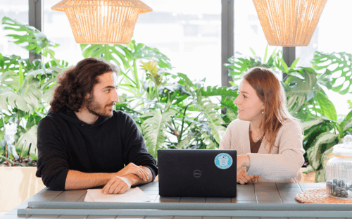 Deux personnes discutent à une table avec un ordinateur portable. L'homme à gauche a les cheveux longs, la femme à droite les cheveux blonds. Plantes vertes en arrière-plan.