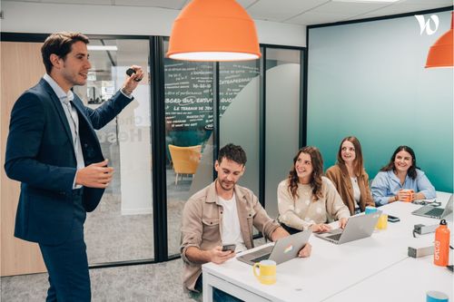 Une femme colle des post-its sur une paroi en verre pendant qu’un homme assis à une table avec un ordinateur portable l’observe. Ils sont dans un bureau lumineux, en pleine séance de travail ou de brainstorming.