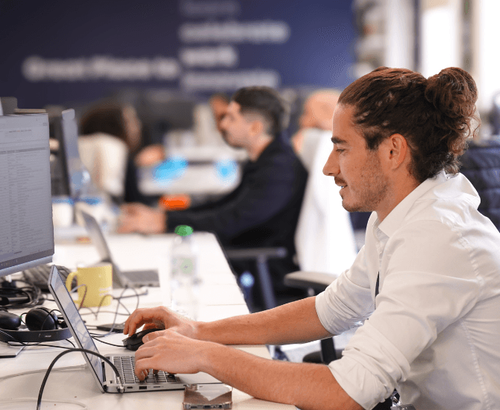 Un homme aux cheveux longs attachés en chignon est assis à un bureau, concentré sur son ordinateur portable et son écran d'ordinateur. D'autres personnes travaillent à des bureaux similaires en arrière-plan dans un espace de bureau ouvert.