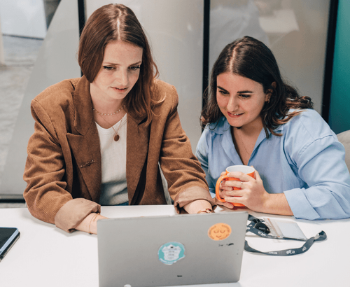 Deux femmes sont assises devant un ordinateur portable. La femme à gauche, aux cheveux bruns, regarde attentivement l'écran. La femme à droite, aux cheveux bruns et à la chemise bleue, tient une tasse orange et regarde également l'écran avec un sourire.
