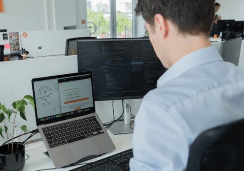Homme de dos travaillant sur deux écrans dans un bureau. L’écran principal affiche du code, tandis que l’ordinateur portable montre un schéma technique et un document professionnel. Une plante verte est posée sur le bureau.