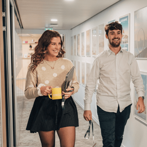 Un homme et une femme souriants marchent dans un couloir de bureau. La femme, au premier plan, tient une tasse jaune et un ordinateur portable.