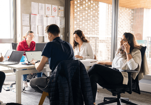 Quatre personnes travaillent autour d’une table dans un bureau lumineux. Deux utilisent un ordinateur portable, une prend des notes et une parle au téléphone. Fenêtres vitrées et affiches au mur en arrière-plan.