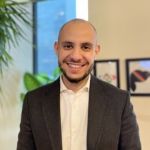 Portrait de Marouane, un homme souriant à la barbe courte, portant une veste sombre sur une chemise blanche. Il se tient dans un bureau avec une plante verte et des cadres décoratifs en arrière-plan.