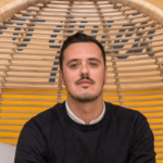 Retrato profesional de Carl, un hombre con bigote, vestido con un jersey negro sobre una camisa blanca. Está sentado en un sillón de mimbre colgante, con un fondo amarillo.