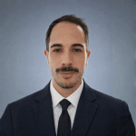 Retrato profesional de Kévin, un hombre con bigote, vestido con un traje azul marino, una camisa blanca y una corbata negra. Posó de frente contra un fondo azul grisáceo neutro.