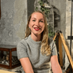 Retrato de Marie, una mujer sonriente de cabello rubio, vestida con un jersey de manga corta gris. Está en un interior de estilo industrial, con una jaula de pájaros blanca y un trípode de madera en el fondo.