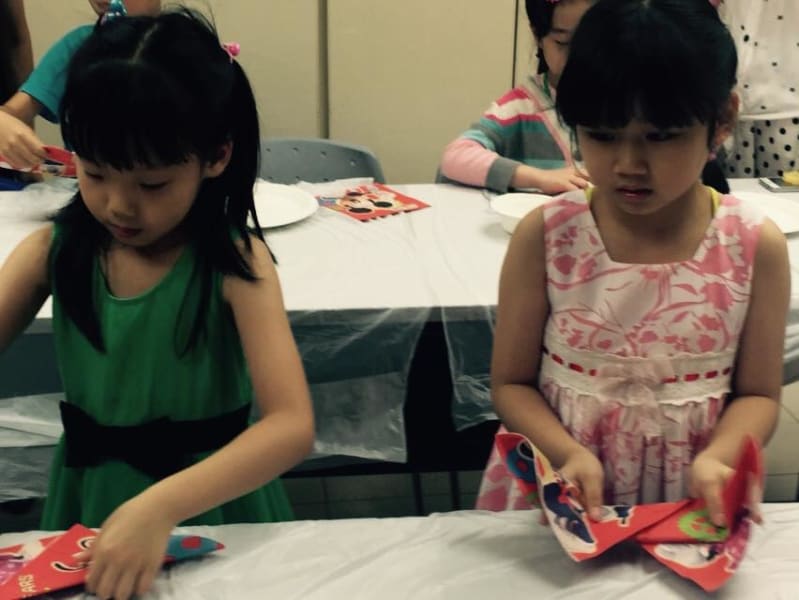 Two young girls at a table, focused on a craft activity with red paper items. One wears a green dress, the other pink.