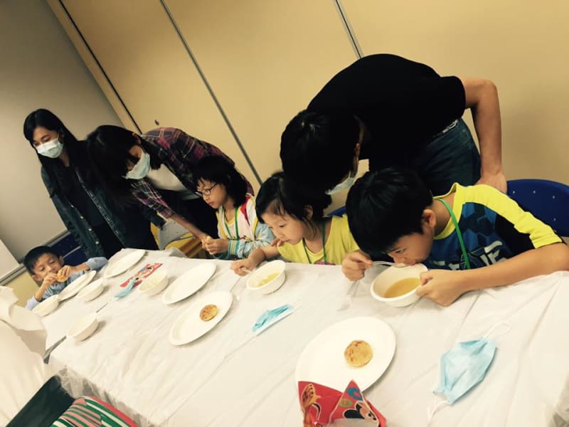 Children and adults at a long table. Three children are eating soup from bowls, while adults supervise them.