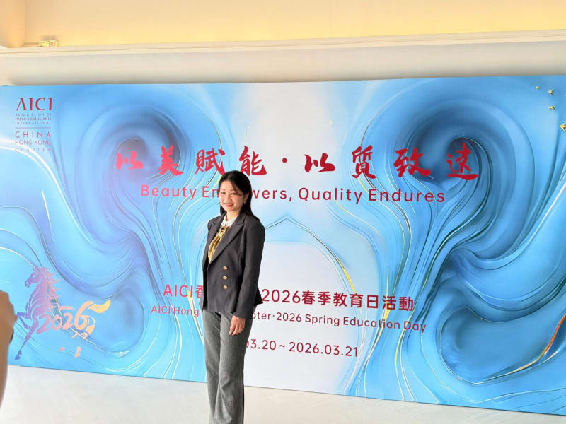 Woman in a dark suit and patterned blouse stands smiling in front of a blue event banner with text.