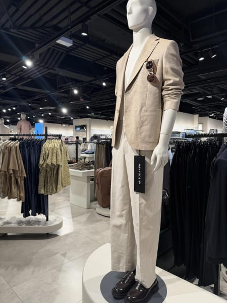 A mannequin wears a beige suit with sunglasses in the jacket pocket and dark loafers, displayed in a clothing store.