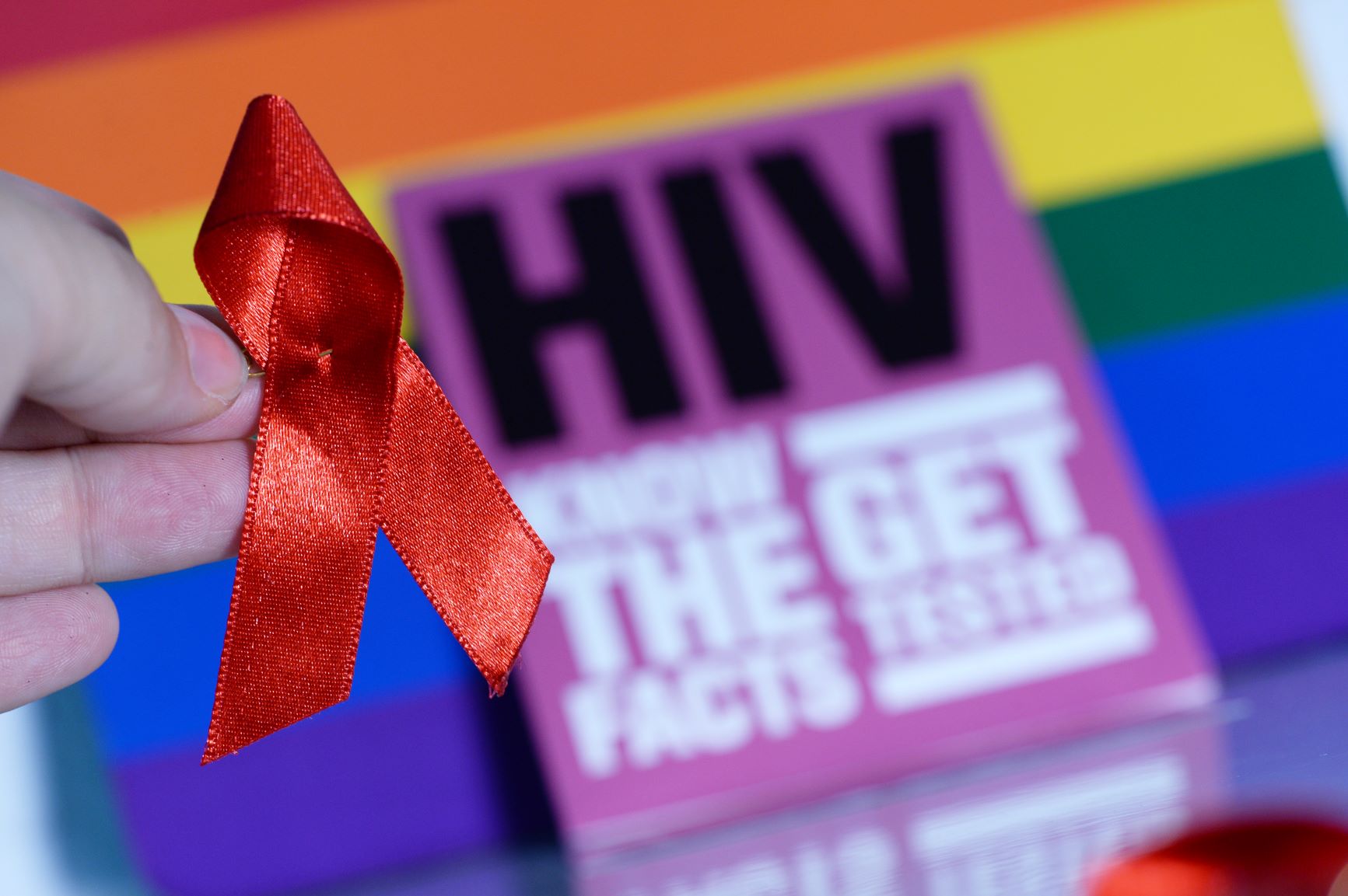 A red HIV ribbon is held in front of a base which is rainbow-coloured A red HIV ribbon is held in front of a base which is rainbow-coloured