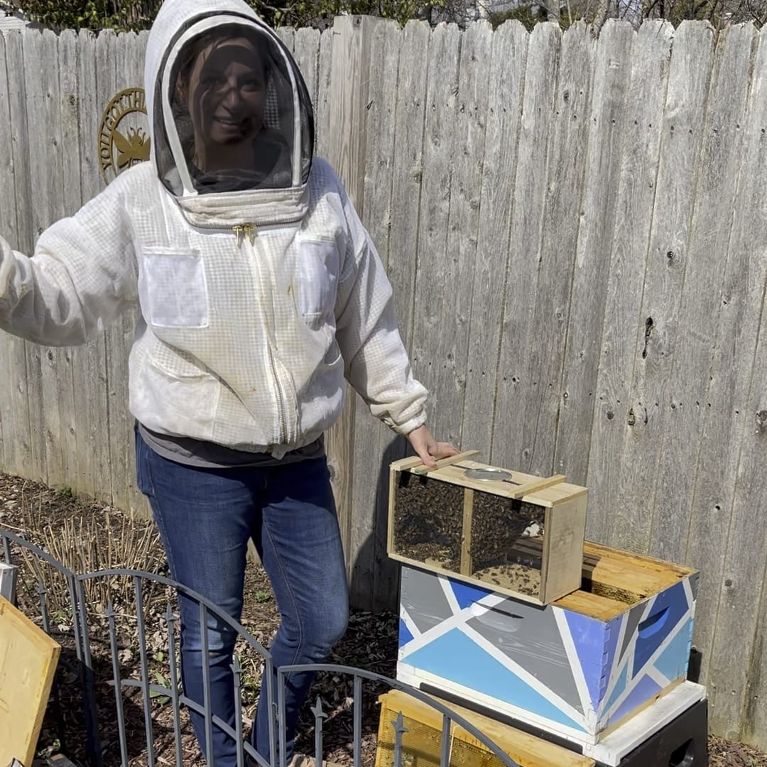Kate in a beekeeper's suit holding a wooden frame with bees, standing beside beekeeping boxes in a garden.