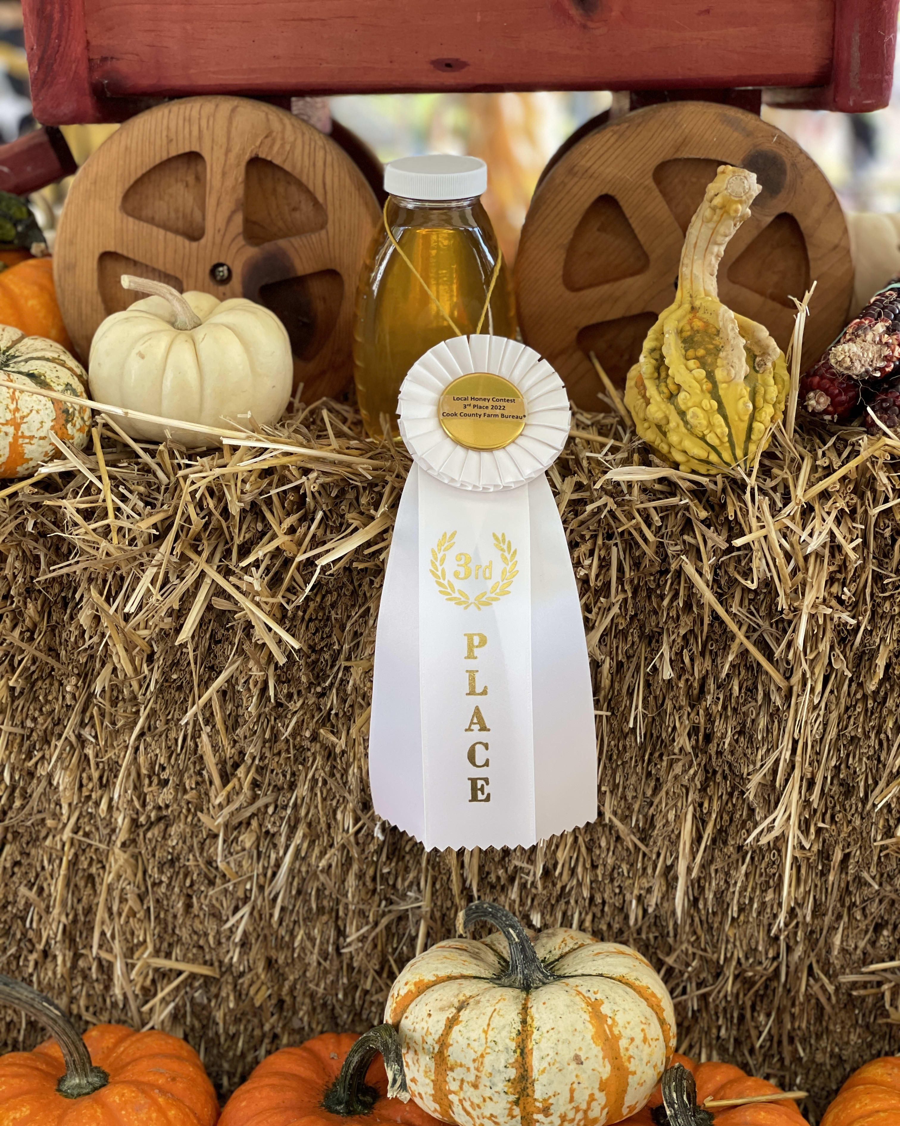 A bottle of honey with a white '3rd place' ribbon, sitting on a hay bale in front of a wooden wagon and some gourds.
